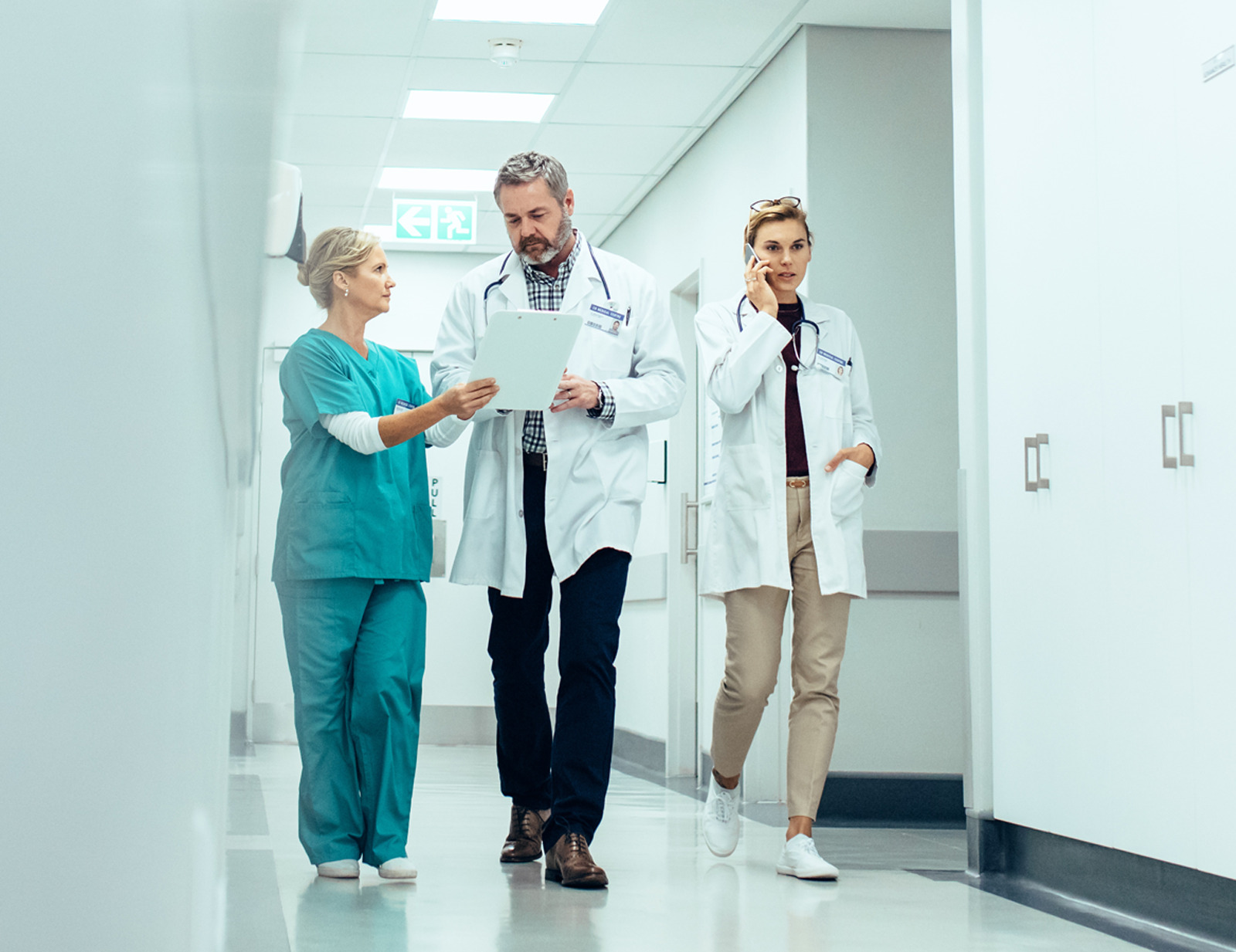 Two doctors and a nurse walking down a corridor, looking at a clipboard and talking on the phone.